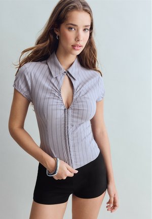 Jeune femme aux longs cheveux bruns portant un haut rayé gris à manches courtes et un short noir, posant avec une main dans la poche sur un fond uni.