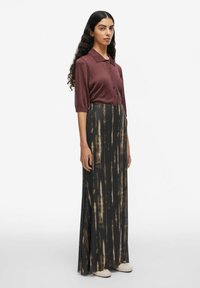 Donna con lunghi capelli scuri, che indossa una camicia bordeaux con bottoni, una lunga gonna tie-dye nera e marrone, e scarpe bianche, in piedi contro uno sfondo bianco.
