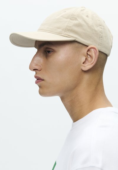 Νέος άνδρας με μπεζ καπέλο μπέιζμπολ και λευκό πουκάμισο, στραμμένος προς τα αριστερά, σε απλό λευκό φόντο.