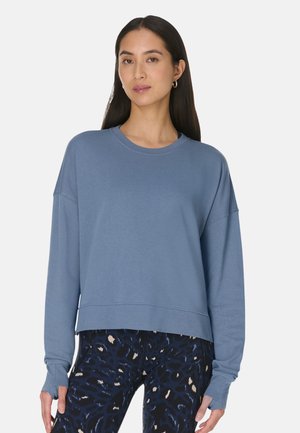 Femme portant un sweat-shirt oversize bleu à manches longues et des leggings sombres à motifs, debout devant un fond clair uni.
