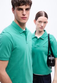 Νέος άνδρας και γυναίκα που φορούν ταιριαστά πράσινα πόλο μπλουζάκια, στέκονται κοντά με ουδέτερες εκφράσεις σε απλό λευκό φόντο.