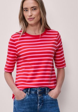 Mujer con cabello rubio largo, vistiendo una camiseta de rayas rojas y rosas de media manga y jeans azules, con las manos en los bolsillos, sobre un fondo liso.