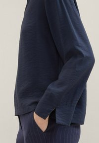 Chemise à manches longues bleu marine en tissu doux et texturé. Présente une coupe décontractée, des fentes latérales et un design simple. Associée à un pantalon à rayures bleues.