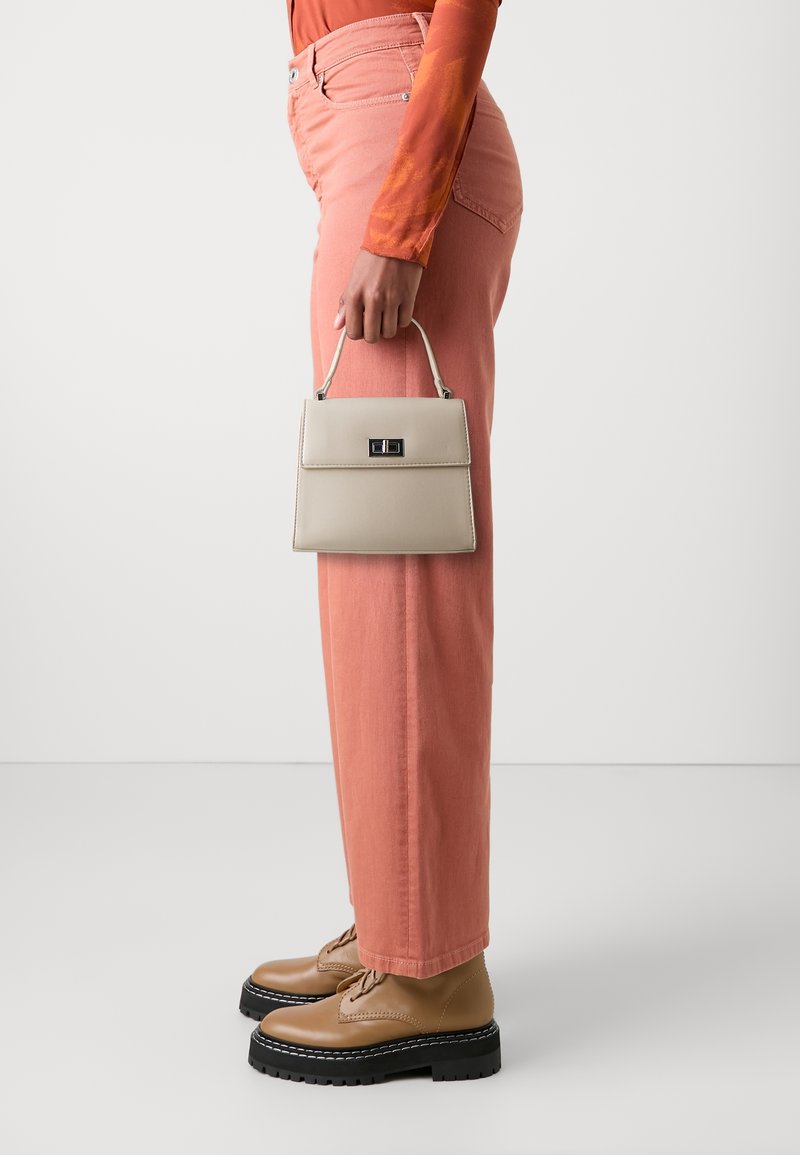 Beiget strukturierte Handtasche mit einem oberen Griff und einem vorderen Verschluss, gehalten in der Hand einer Person, kombiniert mit pinkfarbenen Hosen und braunen schnürschuhen.