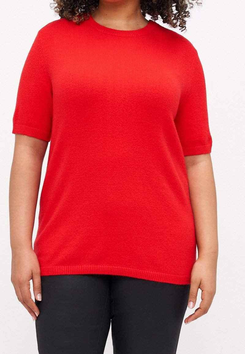 Personne portant un pull tricoté rouge vif à manches courtes et un pantalon noir, avec des cheveux bouclés visibles en haut.