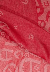 Le tissu rouge présente une texture douce avec un motif répétitif de logos blancs. Le design est léger, mettant en valeur un subtil mélange de couleurs.