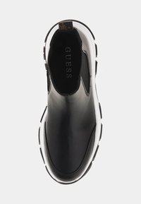Scarpa slip-on in pelle nera con suola in gomma bianca, caratterizzata da una punta arrotondata e un design minimalista. Logo "GUESS" visibile all'interno.
