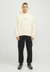 Cream fleece hoodie with a logo, paired with black cargo pants featuring side pockets and black athletic shoes with white soles.