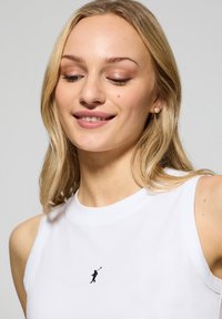 Vit ärmlös topp i bomull med en liten svart broderad figur som håller en golfklubba på bröstet. Enkel och minimalistisk design.