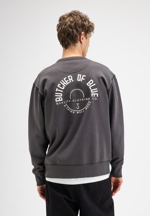 Junger Mann mit lockigem Haar trägt einen dunkelgrauen Sweatshirt mit weißem "Butcher of Blue" Logo und Text auf dem Rücken vor hellem, einfarbigem Hintergrund.