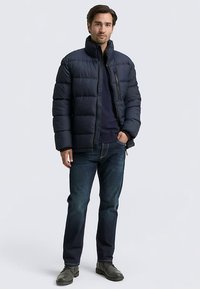 Mörkblå vadderad jacka med horisontellt quiltat mönster, hög krage och dragkedja fram. Bärs över en mörk tröja och kombineras med mörk jeans och stövlar.