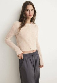 Ljust rosa kort cardigan med rund halsringning, långa ärmar och knappar fram; kombinerad med kolgrå byxor med elastisk midja.