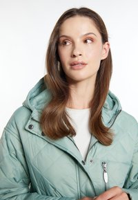 Giacca trapuntata verde menta con cappuccio, dotata di zip frontale, bottoni a pressione e leggera lucentezza. Sotto, un sottogiacca bianco.