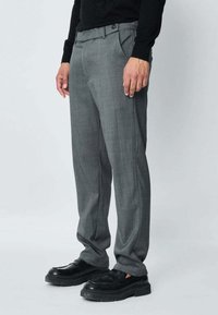 Pantalon gris sur mesure en tissu texturé, avec deux poches avant et une fermeture à bouton unique. Associé à des chaussures à plateforme noires.
