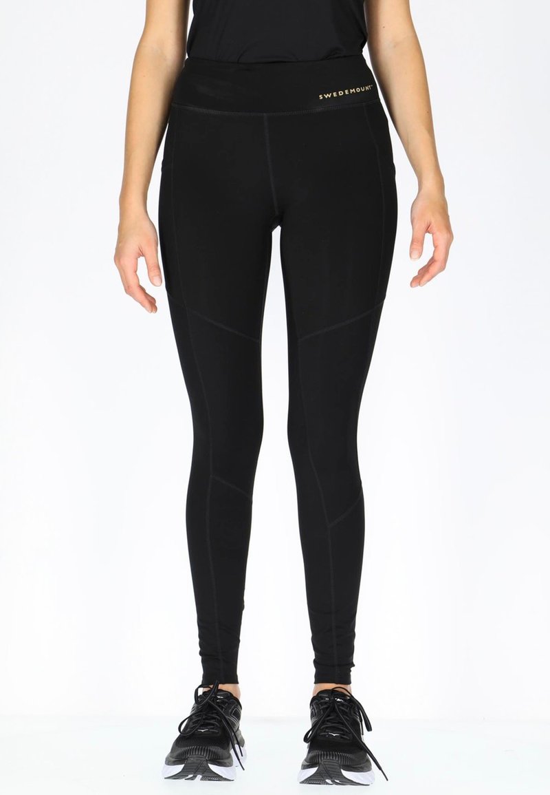 Zwarte leggings met een hoge tailleband, voorzien van contrasterende gestikte naden en een logo op de tailleband. Gecombineerd met zwarte sportieve schoenen.