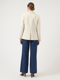 Kreemja tekstuuriga bleiser seljaosaga, kombineeritud laiade tumedate teksapükstega ja beežide kontsadega, vaadatuna tagantpoolt.