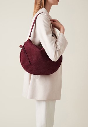 Burgundy textured shoulder bag with a curved shape, featuring a single strap and gold hardware accents. Worn against a light-colored coat.