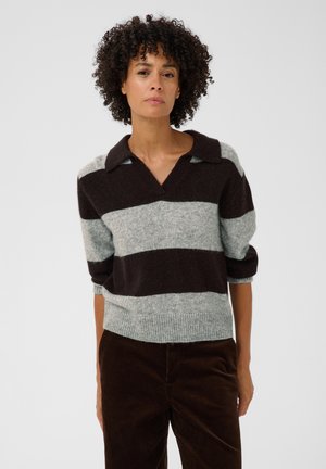 Femme aux cheveux bouclés portant un pull rayé brun foncé et gris clair avec les manches retroussées et un pantalon brun foncé sur un fond uni.
