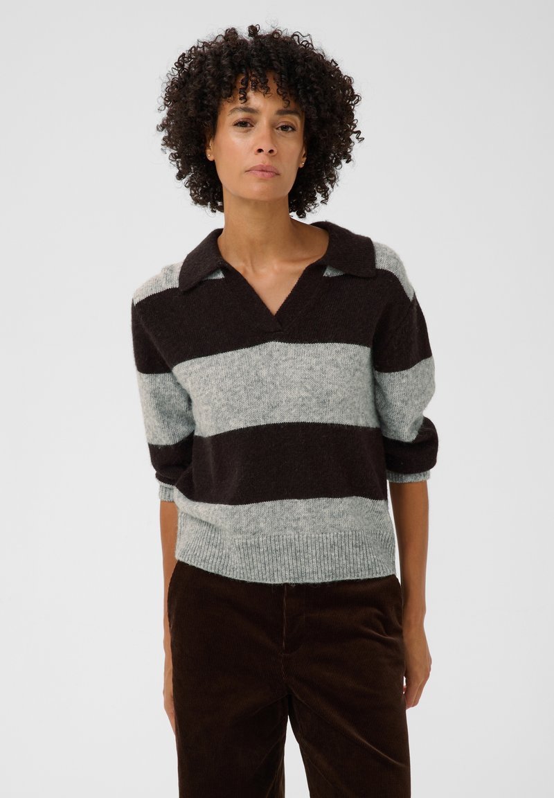Femme aux cheveux bouclés portant un pull rayé brun foncé et gris clair avec les manches retroussées et un pantalon brun foncé sur un fond uni.