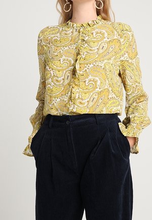 Vrouw draagt een geel paisley-blouse met ruches, gestopt in een zwarte fluwelen broek met hoge taille, met haar handen in de zakken.