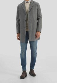 Manteau en mélange de laine grise avec une coupe ajustée, revers crantés et trois boutons, porté sur un pull beige et un jean bleu. Bottines marron.