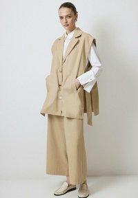 Beige liinasest vest terava kraega, suurte taskute ja kahe nööpiga, millele on sobitatud laiad, sama värvi püksid ja beežid mokassiinid.