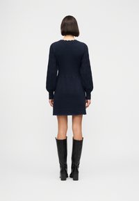 Γυναίκα σε όρθια θέση με την πλάτη γυρισμένη, φοράει ένα ναυτικό πλεκτό φόρεμα με μακριά υφασμένα μανίκια και μαύρες μπότες μέχρι το γόνατο με τακούνι.