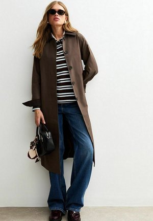 Manteau long marron avec des fermetures à bouton, chemise rayée, jeans bleu à jambe large et un sac à main noir. Le mannequin porte des lunettes de soleil sombres et des chaussures à talons bas.
