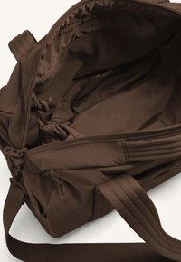 Hnědá nylonová taška uvnitř s hladkým povrchem, se zipovým uzávěrem, síťovou kapsou a prošívanými detaily na bocích a popruzích.