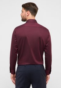 Bordeauxrode blouse met lange mouwen en een gladde textuur, voorzien van een standaardkraag en zwarte knoopaccenten op de manchetten, gezien vanaf de achterkant.