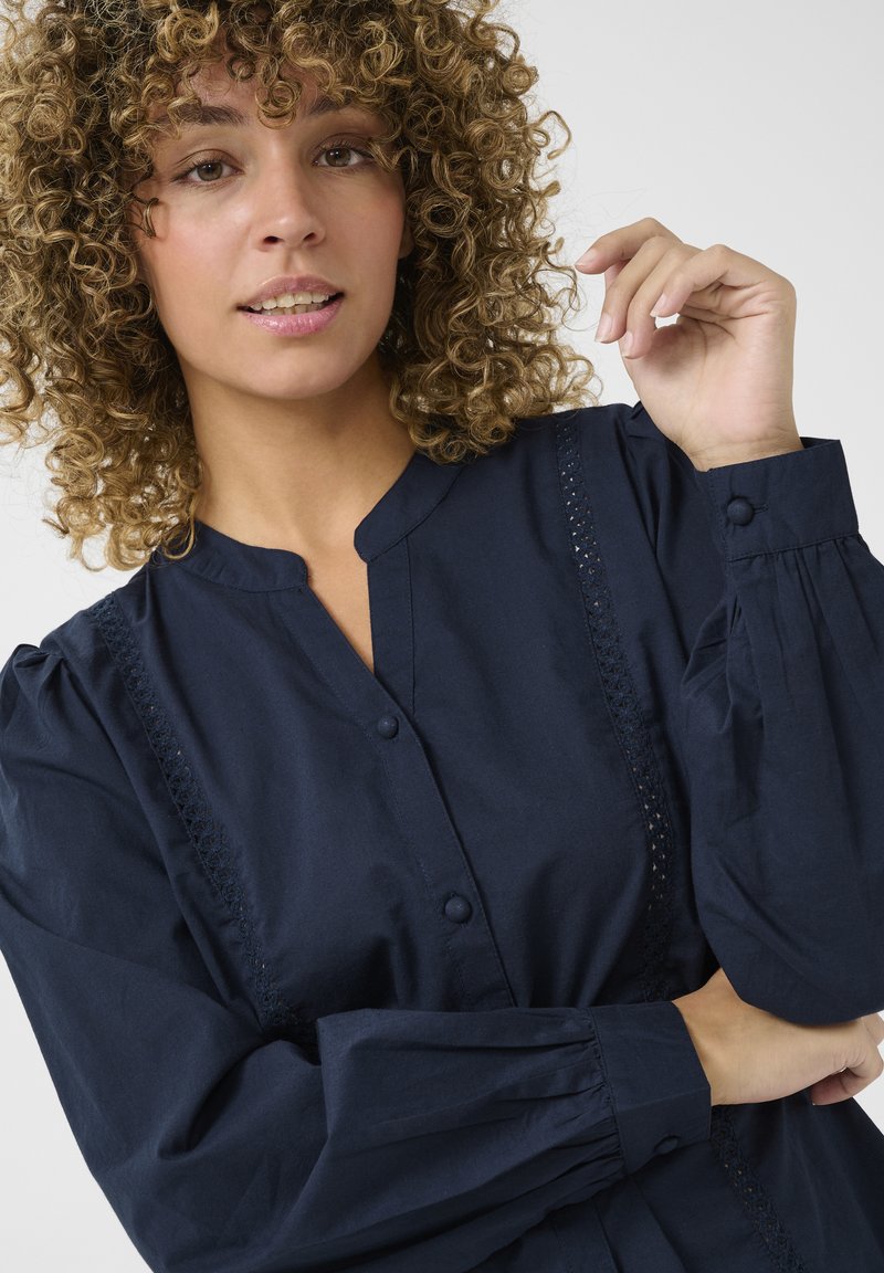 Navyblauwe katoenen blouse met een knoopsluiting aan de voorkant, geplooide mouwen en kantdetails langs de zijkanten. Eenvoudig, gestructureerd ontwerp.