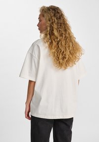 Crème-kleurige oversized katoenen t-shirt met korte mouwen en ronde halslijn, gedragen met donkere broeken, gezien vanuit de achterkant.