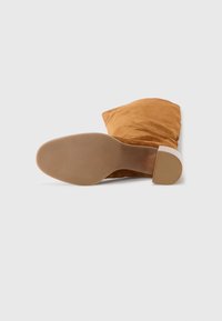 Bottine en suede marron avec une texture douce, un talon cylindrique et une semelle beige lisse, mettant en valeur une pointe arrondie et un design loose.