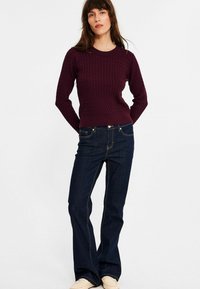 Donker bordeaux gebreid sweater met een gestructureerd ruitpatroon, ronde hals en lange mouwen, gecombineerd met donkerblauwe flared jeans en lichte schoenen.