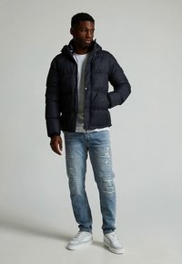 Zwarte donsjas met kap, met een gestructureerd patroon. Gecombineerd met lichtblauwe gescheurde jeans en witte sneakers.