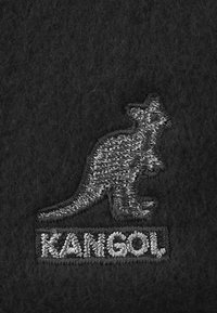Logo di canguro ricamato in filo bianco su tessuto nero con la parola "KANGOL" sotto il canguro in lettere cucite bianche.