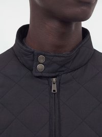 Gros plan d'une personne portant une veste matelassée noire avec un col montant, une fermeture éclair et deux boutons-pression sur le col.
