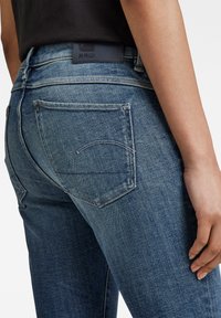 Jeans de denim de color azul oscuro con un diseño clásico de cinco bolsillos, costuras en contraste y una etiqueta de cuero con la marca "RAW" en la cinturilla.