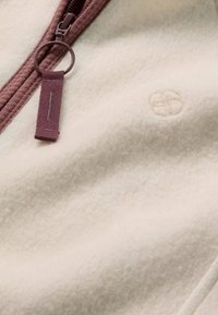 Tissu polaire de couleur crème avec une fermeture éclair mauve et un anneau rond comme tirette, comprenant un petit détail de fleur cousue à côté de la fermeture éclair.