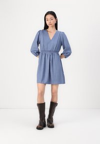 Modra chambray obleka z globokim V-izrezom, napihnjenimi rokavi, elastičnim pasom in obrobnim okrasom, v kombinaciji s svetlobo rjavimi usnjenimi visokimi čevlji do kolen.