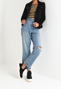 Czarna zamszowa kurtka motocyklowa w połączeniu z paskowanym crop topem, jasnoniebieskimi dżinsami z przetarciami oraz czarnymi trampkami z białą podeszwą.