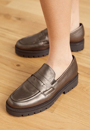 Mocassins en cuir bronze avec une finition texturée, présentant un détail de sangle et une épaisse semelle en caoutchouc noir. Design traditionnel avec une touche moderne.