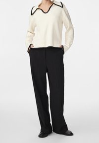 Weißer Pullover mit schwarzem gewelltem Saum am Halsausschnitt und am Saum, kombiniert mit lockeren schwarzen Hosen und schwarzen Schuhen. Einfaches, sauberes Design.