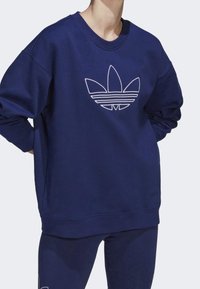 Osoba v námořnickém modrém svetru a kalhotách s velkým bílým konturovým logem Adidas trefoil na hrudi, pózující s rukama za zády.