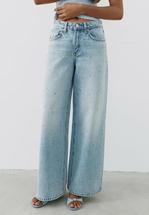Jeans bleu clair à jambes larges avec petits détails en strass bleu, portés avec des sandales ouvertes bleues assorties sur un fond uni.