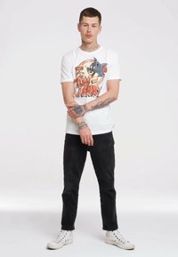 Weißes Baumwoll-T-Shirt mit einem Retro-Tom-und-Jerry-Graphic. Schwarze Denim-Jeans und weiße Sneakers runden den Look ab. Tätowierungen sind auf den Armen sichtbar.