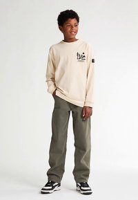 Cămașă crem cu mâneci lungi și design grafic, asortată cu pantaloni verde măsliniu și adidași negri, cu accente albe și un logo pe mânecă.