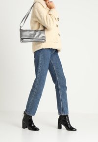 Hopeanvärinen metallinen olkalaukku, jossa on tyylikäs muotoilu, yhdistetty kermaiseen teksturoituun takkiin, sinisiin suoralahkeisiin farkkuihin ja mustiin kiiltonahkaisiin korollisiin saappaisiin.