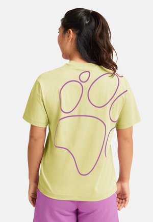 Hellgrünes Baumwoll-T-Shirt mit großem lila abstraktem Design auf der Rückseite. Kurze Ärmel und entspannter Sitz. Kombiniert mit lila Shorts.