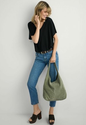 Kobieta w czarnej bluzce z dekoltem w serek i niebieskich dżinsach trzyma oliwkową zamszową torbę typu tote, ma na nogach czarne sandały na platformie, stoi na tle szarej ściany.